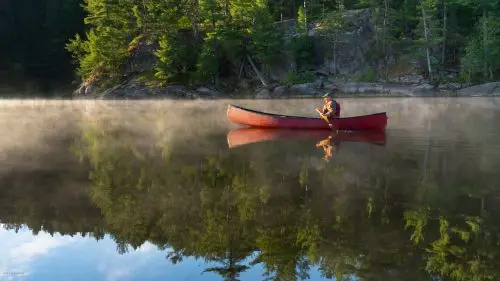 A lone paddler in a red canoe crossing a misty lake at sunrise with forest reflections on calm water.