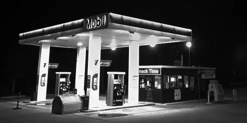 Black-and-white photograph of a brightly lit service station at night with fuel pumps under an illuminated canopy.